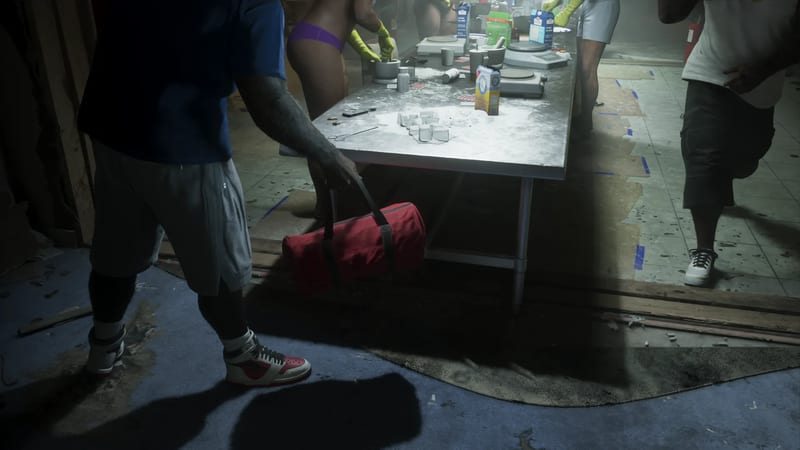 A man in a blue t-shirt and grey shorts carries a red holdall towards a long metal table covered in white powder, with several people working at it.
