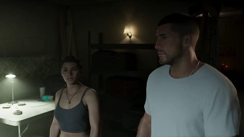 Lucia Caminos and Jason Duval stand in a dimly lit room, she looking ahead, he glancing to the side, beside a table with a lamp.