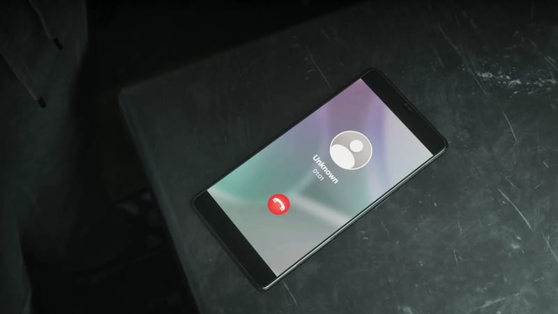 A smartphone lies on a dark, scratched surface showing a call from "Unknown" at 01:01, with a red end-call button on screen.