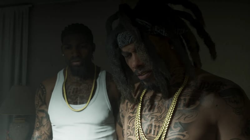Two heavily tattooed men in gold chains stand in a dim room, one in a white vest, the other shirtless with long dreadlocks and a bandana.
