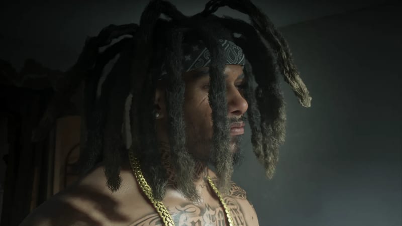 A shirtless, tattooed man with long dreadlocks, a patterned bandana and a thick gold chain looks to the right in a dim, smoky room.