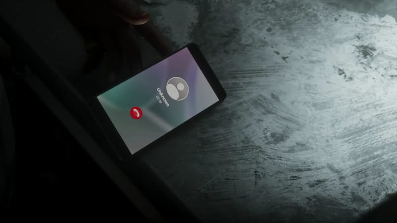 A smartphone lies on a scratched, pale grey surface in a dark room, its screen showing a call from "Unknown" with a red end-call button.
