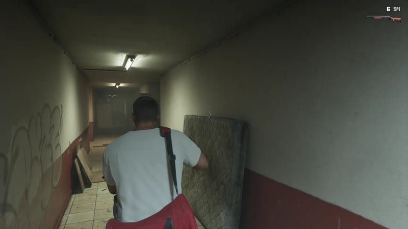 A man in a white T-shirt with a red bag walks down a dim, grubby corridor past a stained mattress propped against the wall.