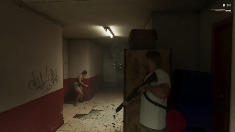 A man in a white T-shirt aims a shotgun from behind a board while a woman with a pistol crouches against the wall of a dim red and cream corridor.