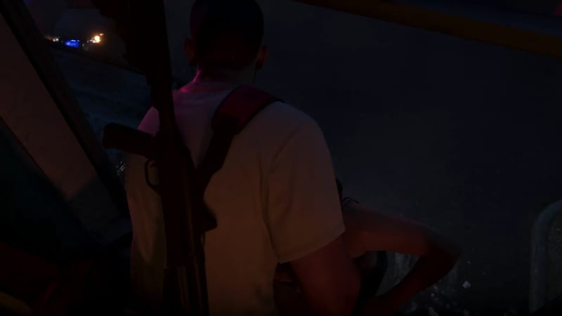 A man in a white T-shirt with a rifle slung over his shoulder is seen from behind in a dark, dimly lit space at night.