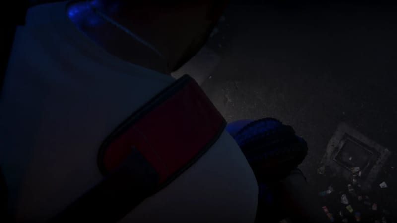 A very dark, high-angle view of a person in a white top with a red-trimmed strap, beside another person with braided hair, on a littered night-time pavement.