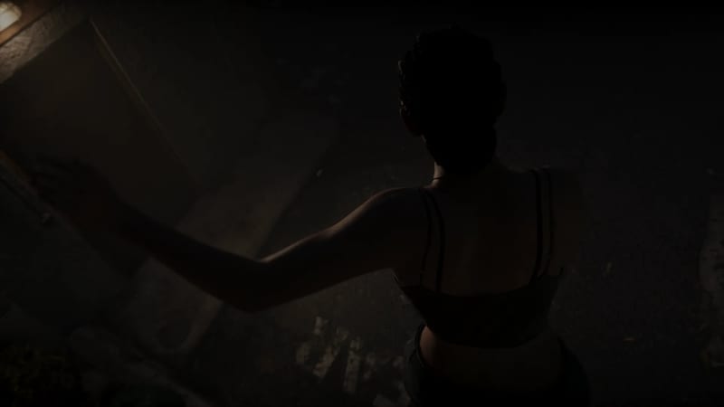 A woman seen from behind in near darkness, one arm stretched out to her left, wearing a strappy top, with a dim light glowing at the top left corner.