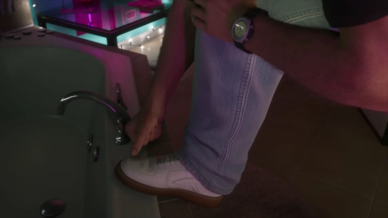 A man with a purple wristwatch ties the laces of a white trainer, his foot resting on the edge of a bathtub in a dimly lit, neon-pink bathroom.