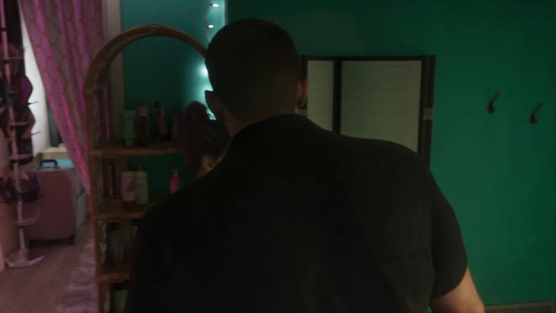 A broad-shouldered man in a dark shirt, seen from behind, stands in a dim teal bedroom beside a cane shelf, with a woman partly hidden beyond him.