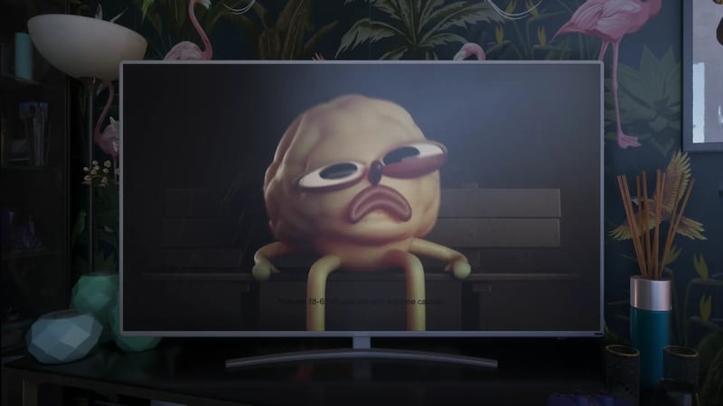 A cartoon blob character with a sad face sits on a bench on a widescreen TV in a dim room with flamingo wallpaper.