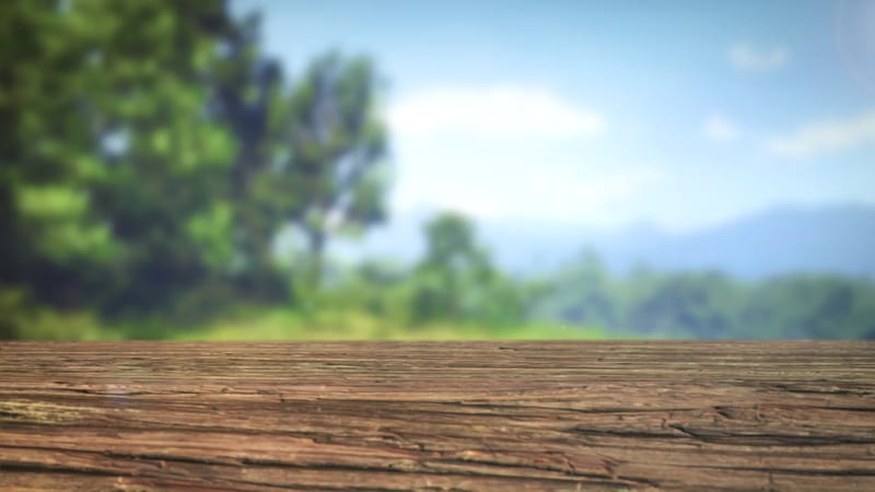 A weathered wooden surface in sharp focus in the foreground, with blurred green trees, grassy hills and a hazy blue sky behind.