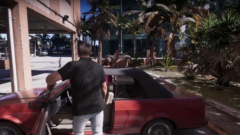 A man in a black short-sleeved shirt and light jeans stands by the open door of a red convertible while a woman sits inside, on a sunny palm-lined street.