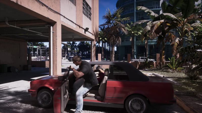 A man in a black shirt and light jeans climbs out of a red convertible with a black roof, parked beside a pink building with palm trees behind.
