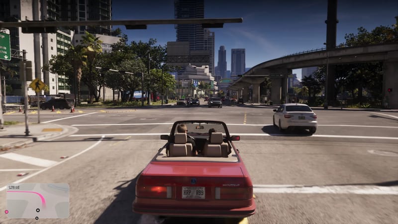 A red convertible with two people in it waits at a wide city junction in Vice City, with tall towers, palm trees and a raised rail track beyond.