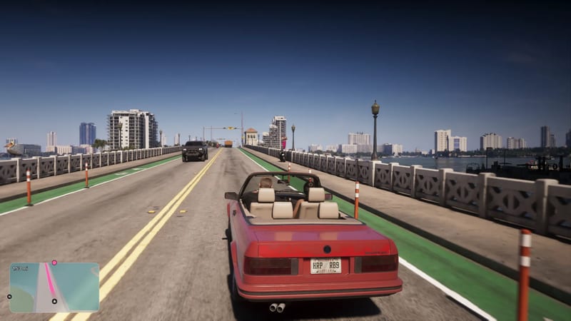 A red convertible with cream seats drives along a bridge in Vice City on a sunny day, with white tower blocks and calm water beyond.