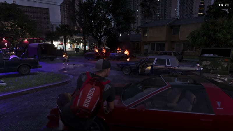 A man in a grey cap and red BIGNESS backpack crouches behind a red car on a night street where two cars burn and a man stands in flames nearby.
