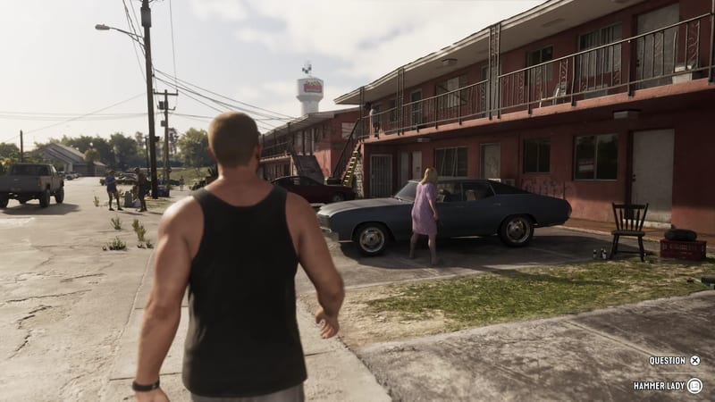 Jason Duval walks along a sunlit motel car park towards a woman in a lilac dress beside a dark blue classic car, with a water tower behind.