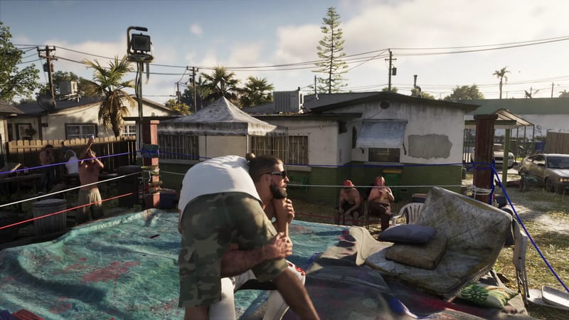 A bearded man in sunglasses and camouflage shorts lifts another man in a backyard wrestling ring while onlookers watch beside small houses.