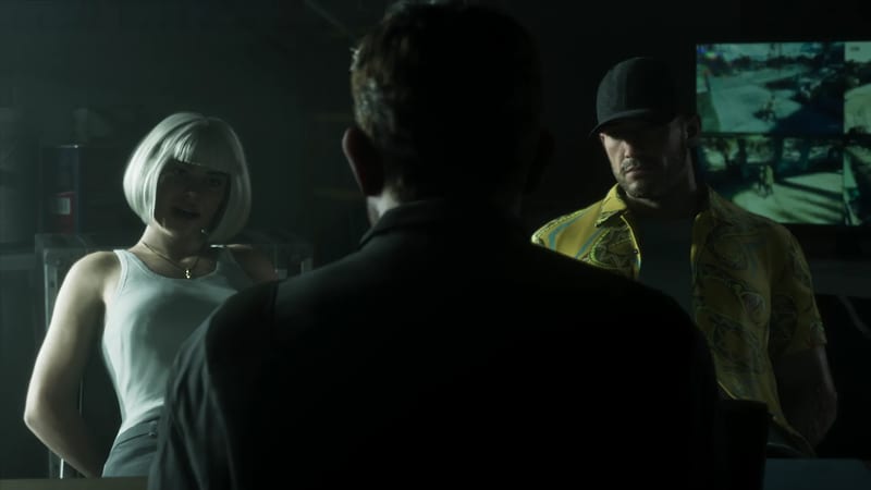 A woman with a white bob and a man in a yellow patterned shirt and black cap face a dark figure seen from behind, in a dim room with a screen.