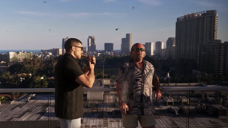 Jason Duval lights a cigar beside Brian Heder on a rooftop terrace, with a city skyline of tower blocks behind them in golden evening light.
