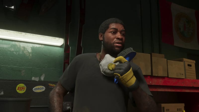 A bearded man in a grey t-shirt and yellow work gloves holds a dust mask and a blue scrub brush in a green-walled workshop.