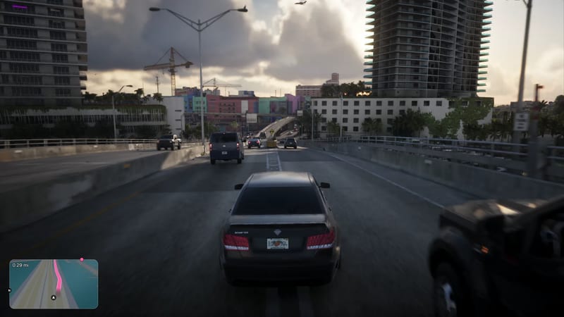 A dark grey Schafter saloon drives along a raised multi-lane road past tower blocks and colourful low buildings under a cloudy sky.