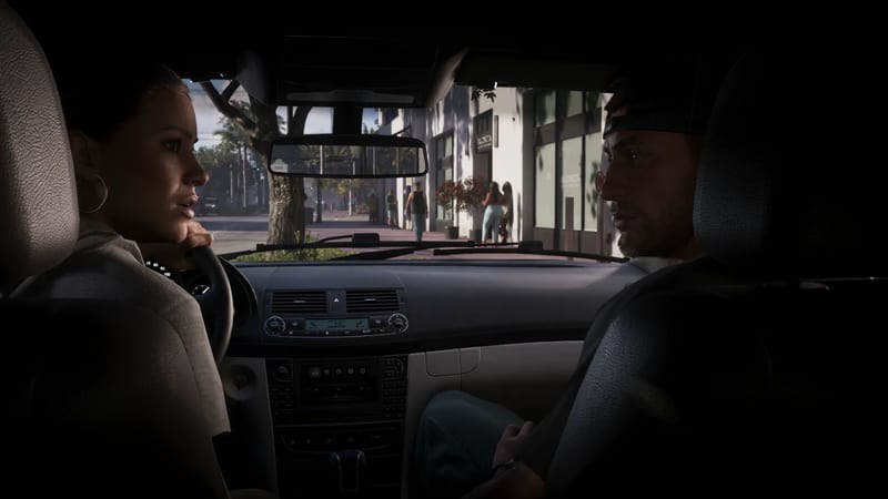 Lucia Caminos and Jason Duval sit in the front of a car, looking at each other, with a sunny city street and pedestrians seen through the windscreen.
