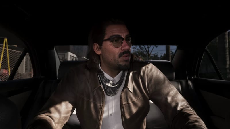 A man with a moustache, goatee and tinted glasses sits in the back of a car, wearing a brown leather jacket and a patterned neckerchief.