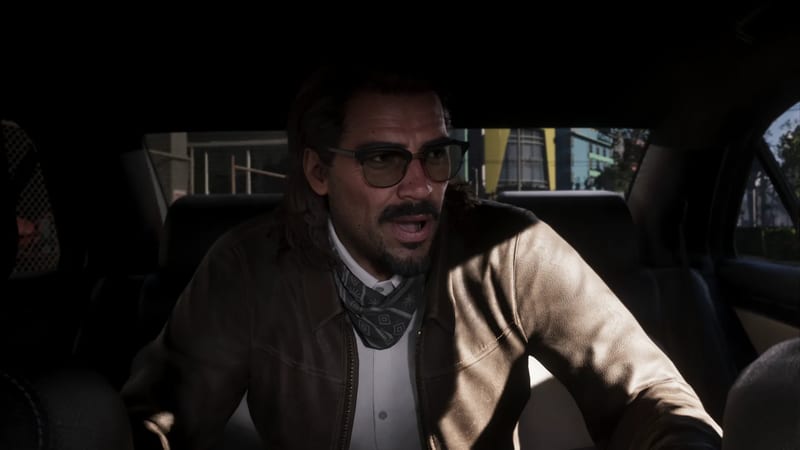 A man with a moustache, glasses and a grey bandana speaks while sitting inside a dim car, with a sunlit street visible through the windows.