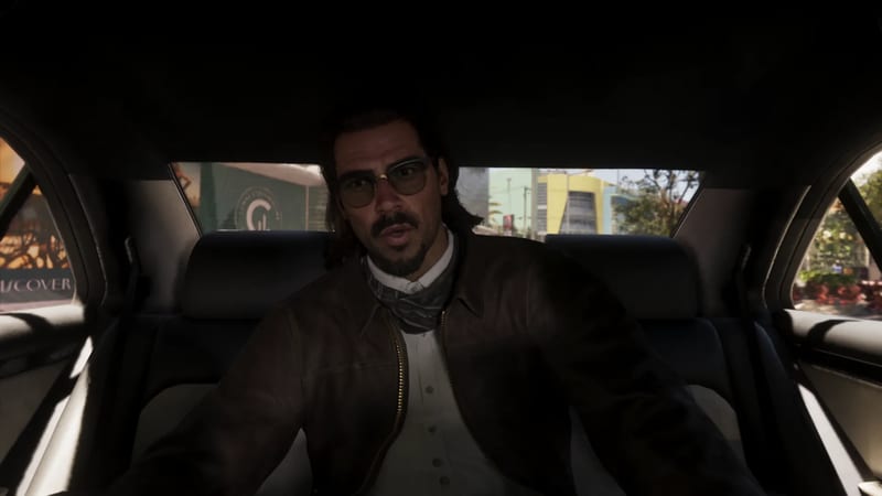 A man with a moustache, goatee and tinted glasses sits in the back seat of a car, with a sunlit street visible through the windows behind him.