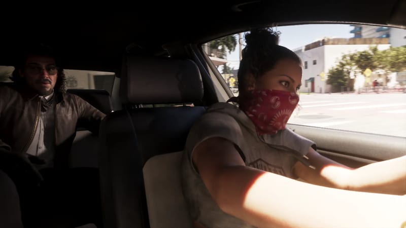 A woman in a red bandana face covering drives a car while a man in tinted glasses sits in the back seat, with a sunlit street outside.