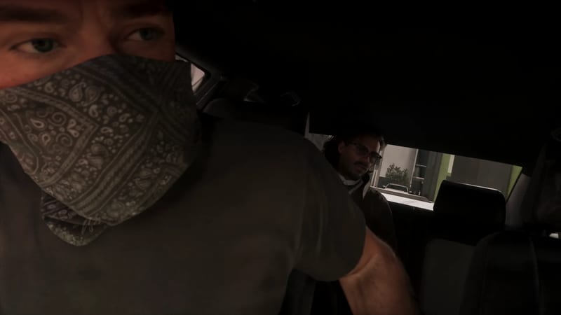 A man in a grey T-shirt and paisley bandana over his face sits in a dark car, while a bearded man in glasses watches from the back seat.
