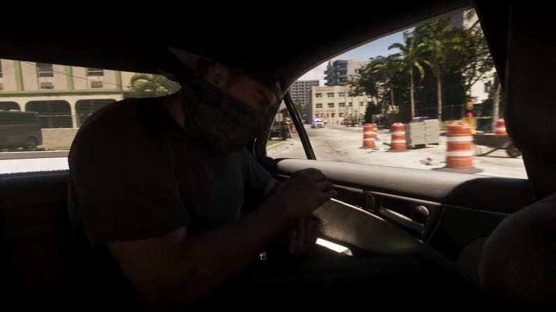 A masked man in a cap and dark top crouches low in a car's back seat, with a police car and traffic barrels visible through the window.