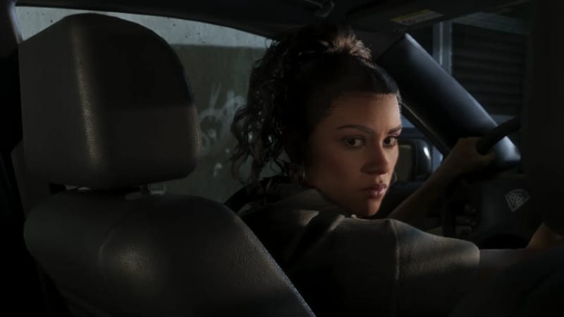 Lucia Caminos sits at the wheel of a car in a dim space, hand on the steering wheel, looking back over her shoulder at the camera.