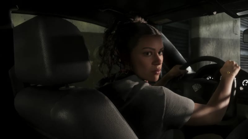 Lucia Caminos looks back over her shoulder from the driver's seat of a car, both hands on the wheel, in a dimly lit concrete space.