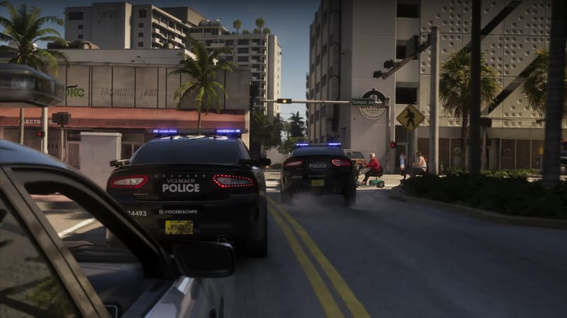 Two black Vice Beach Police cars with blue lights flashing drive down a sunlit city street past an older person on a green mobility scooter.