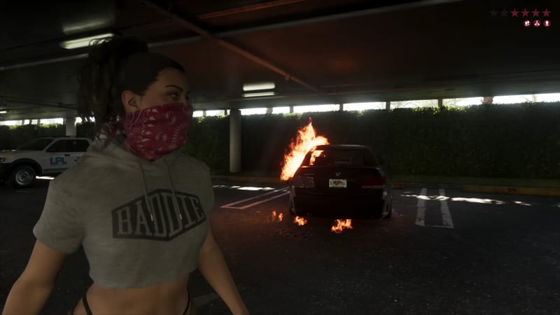 Lucia Caminos, wearing a red bandana over her face and a grey "BADDIE" crop hoodie, stands in a car park near a burning dark saloon car.