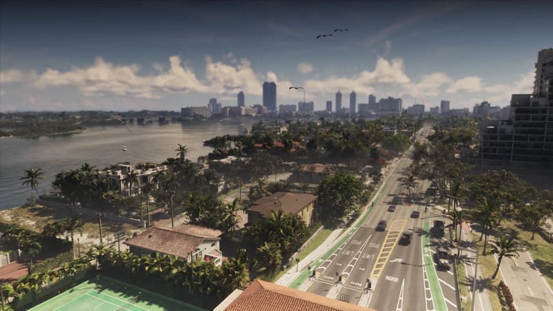 A wide road runs beside a bay lined with palm trees and houses, with a distant city skyline under a blue sky with white clouds in Vice City.
