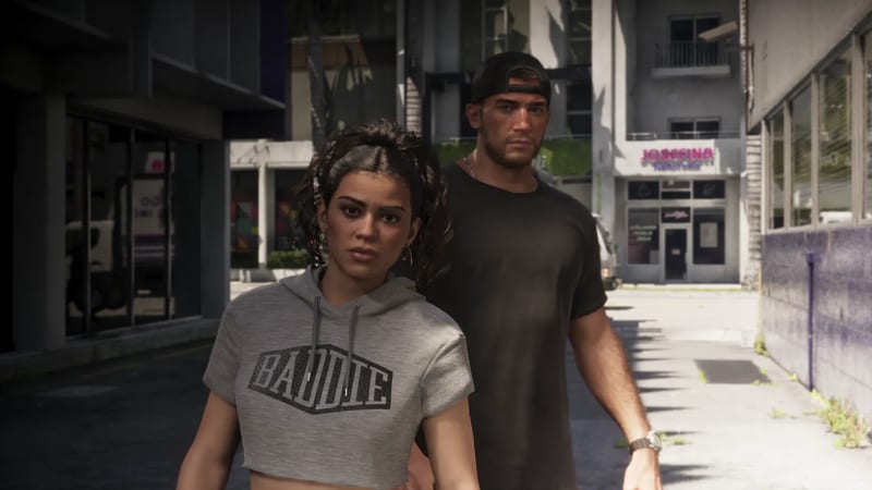 Lucia Caminos in a grey cropped hoodie stands in front of Jason Duval, who wears a backwards cap, on a sunlit city street.