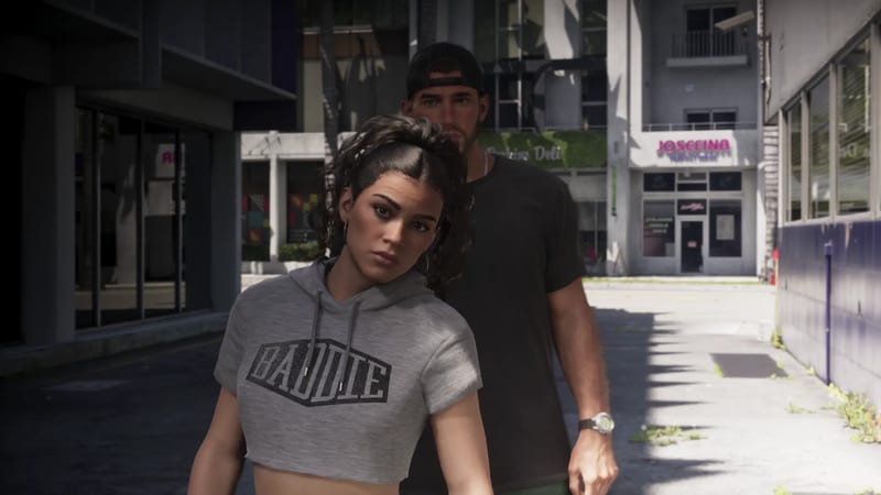 Lucia Caminos, in a grey cropped hoodie, looks warily at the camera as Jason Duval stands behind her on a sunlit city street.