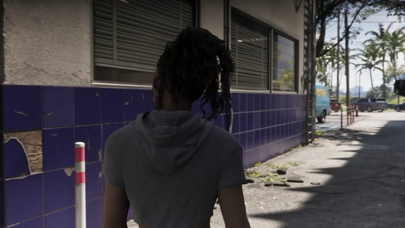A woman with dark curly hair in a grey cropped hoodie walks away from the camera along a sunlit alley beside a building with a purple tiled base.