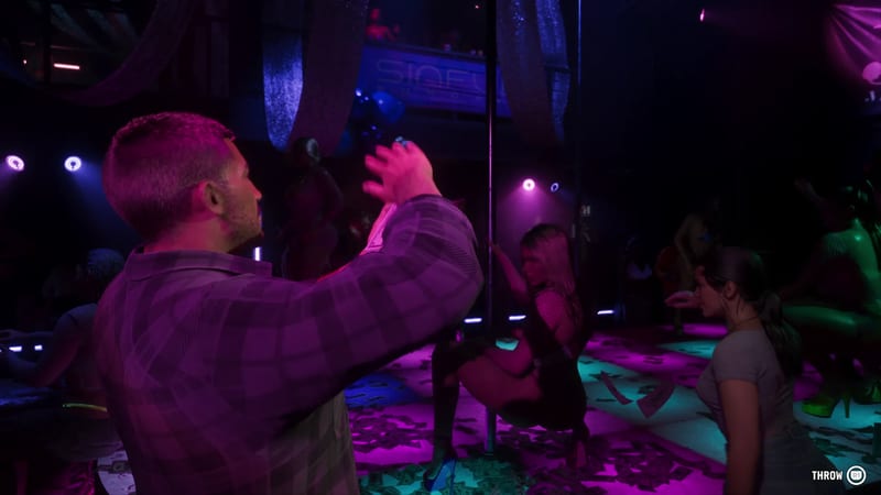 A man in a plaid shirt throws banknotes in a dim, neon-lit club as a woman dances at a pole and another woman watches, with cash scattered on the floor.