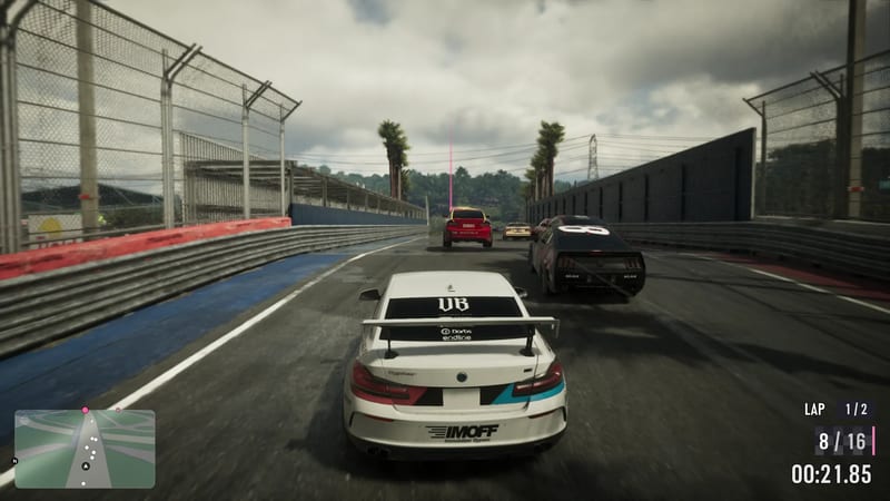 A white racing saloon with a rear wing follows a red car and a black car along a fenced race track under a cloudy sky, with a race display on screen.