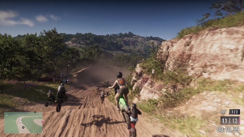 A woman in a black helmet rides a green dirt bike up a dusty track, chasing other riders, with a rocky cliff on the right on a sunny day.
