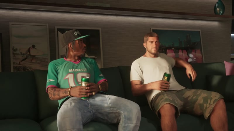 Two men sit on a dark green sofa holding cans of Benedict beer, one in a green Manatees 19 jersey and cap, the other in a white T-shirt and camouflage shorts.