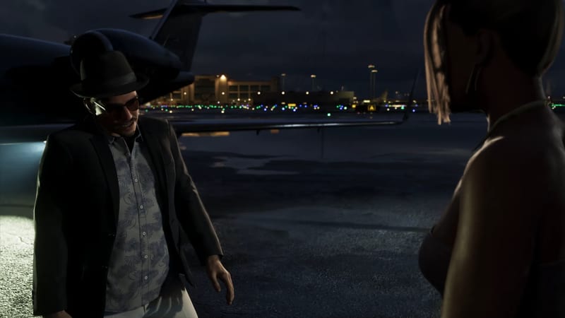 A man in a black fedora, sunglasses and blazer stands beside a dark private jet on an airport apron at night, facing a woman in the foreground.