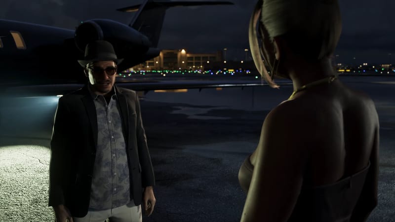 A man in a black hat, sunglasses, dark blazer and patterned shirt talks to a woman seen from behind, beside a dark private jet on an airfield at night.