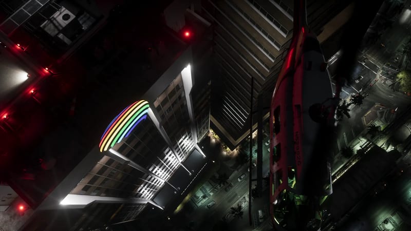A Buckingham Maverick helicopter seen from directly above banks over city rooftops at night, beside a tower with a glowing rainbow sign.