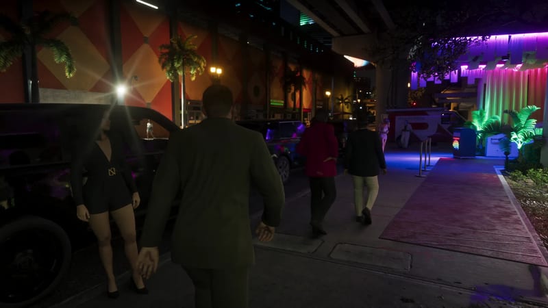A man in a dark green suit, seen from behind, walks along a neon-lit night-time pavement past a woman in black beside a parked dark car.