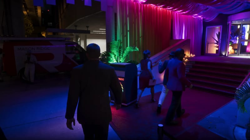 A man in a dark suit walks towards people heading up a red-carpeted entrance lit in pink, purple and green, with a white van nearby at night.